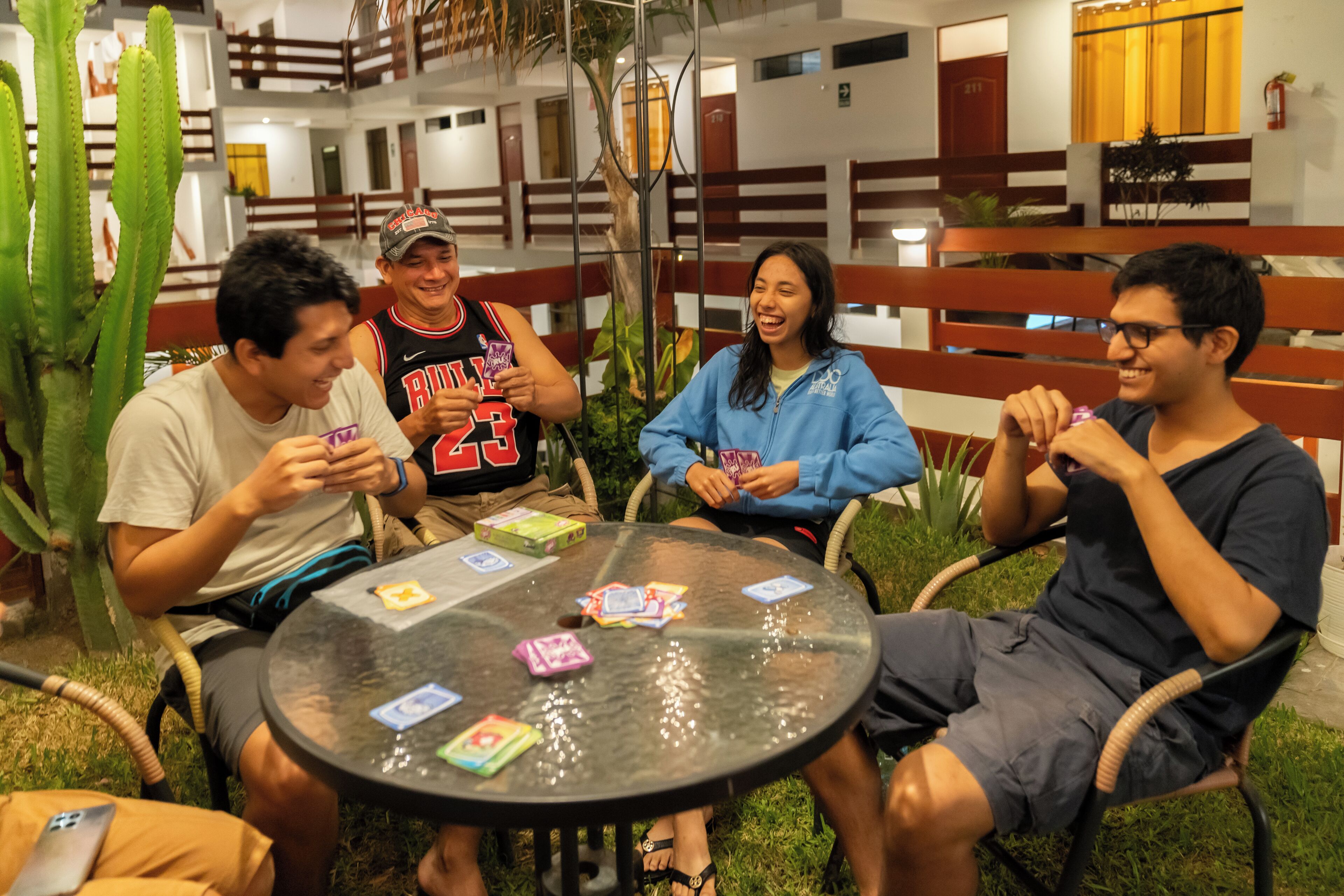Un grupo de personas jugando cartas en una mesa al aire libre con superficie de vidrio, rodeado de vegetación y un edificio con balcón al fondo.