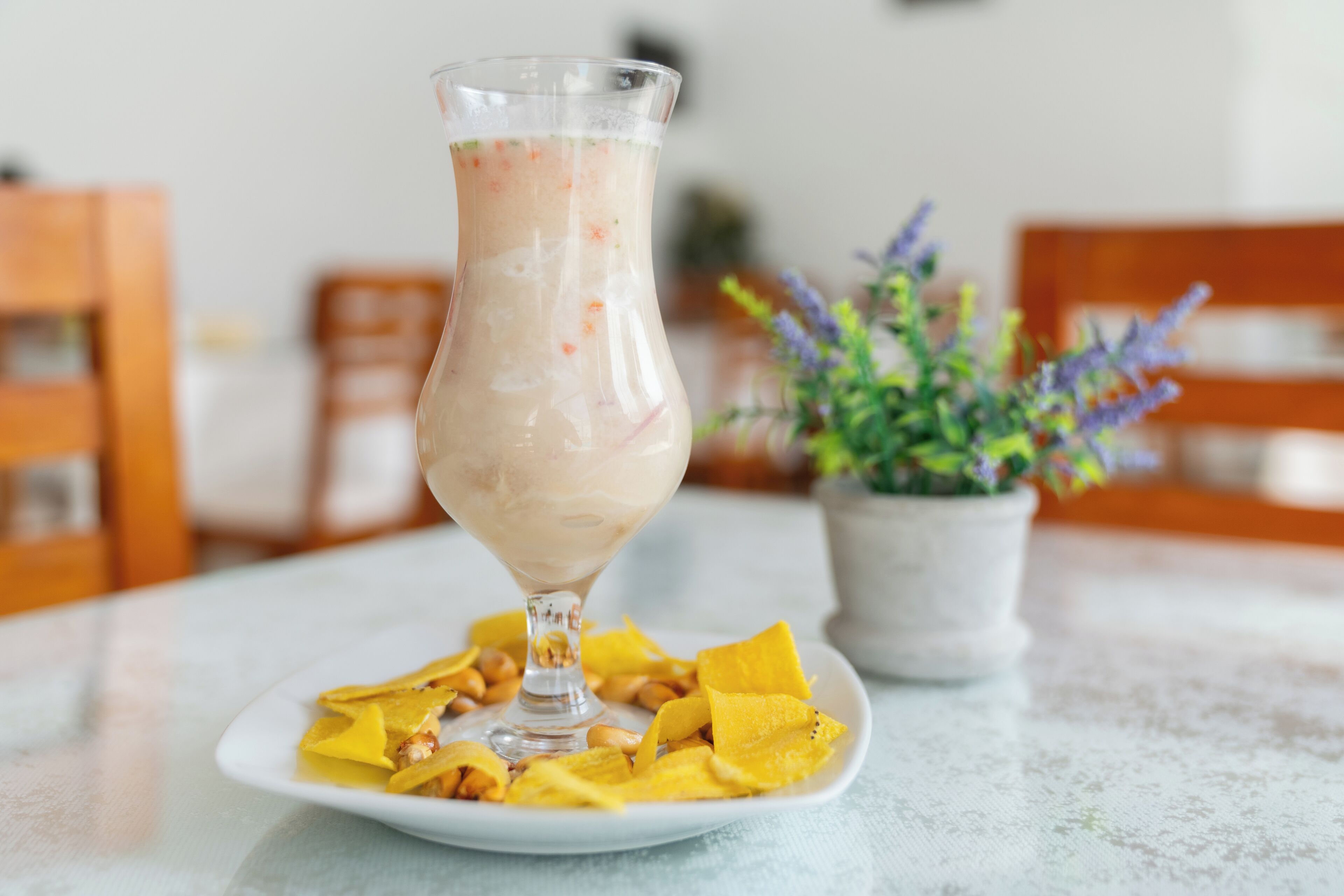 A glass of a creamy beverage on a white plate with yellow fruit pieces, set on a table with a potted plant in the background.