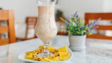 Un vaso de una bebida cremosa sobre un plato blanco con trozos de fruta amarilla, colocado sobre una mesa con una planta en maceta al fondo.