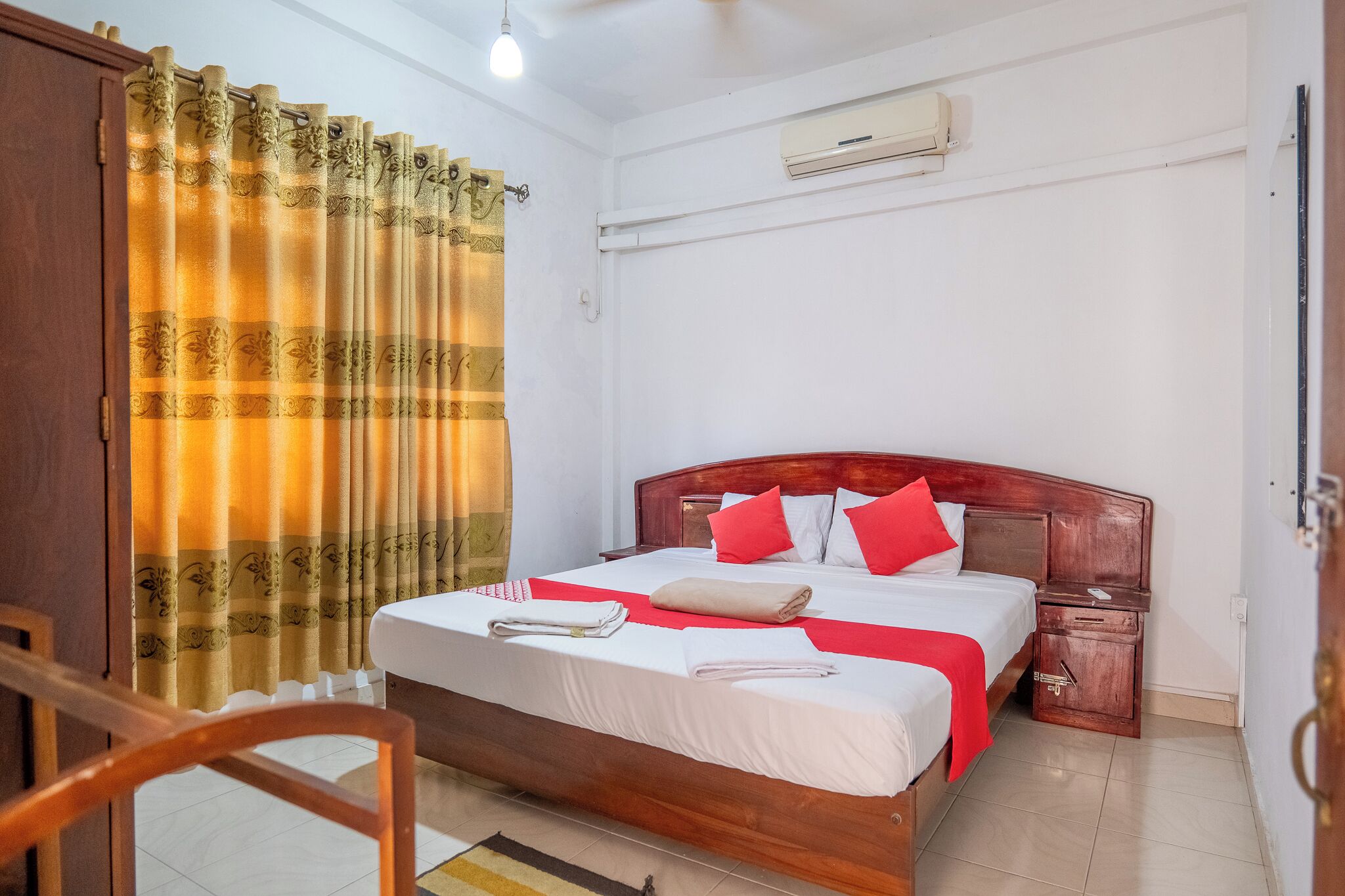 A neatly made bed with red and white bedding, a wooden headboard, a nightstand, and a curtain with a floral pattern.
