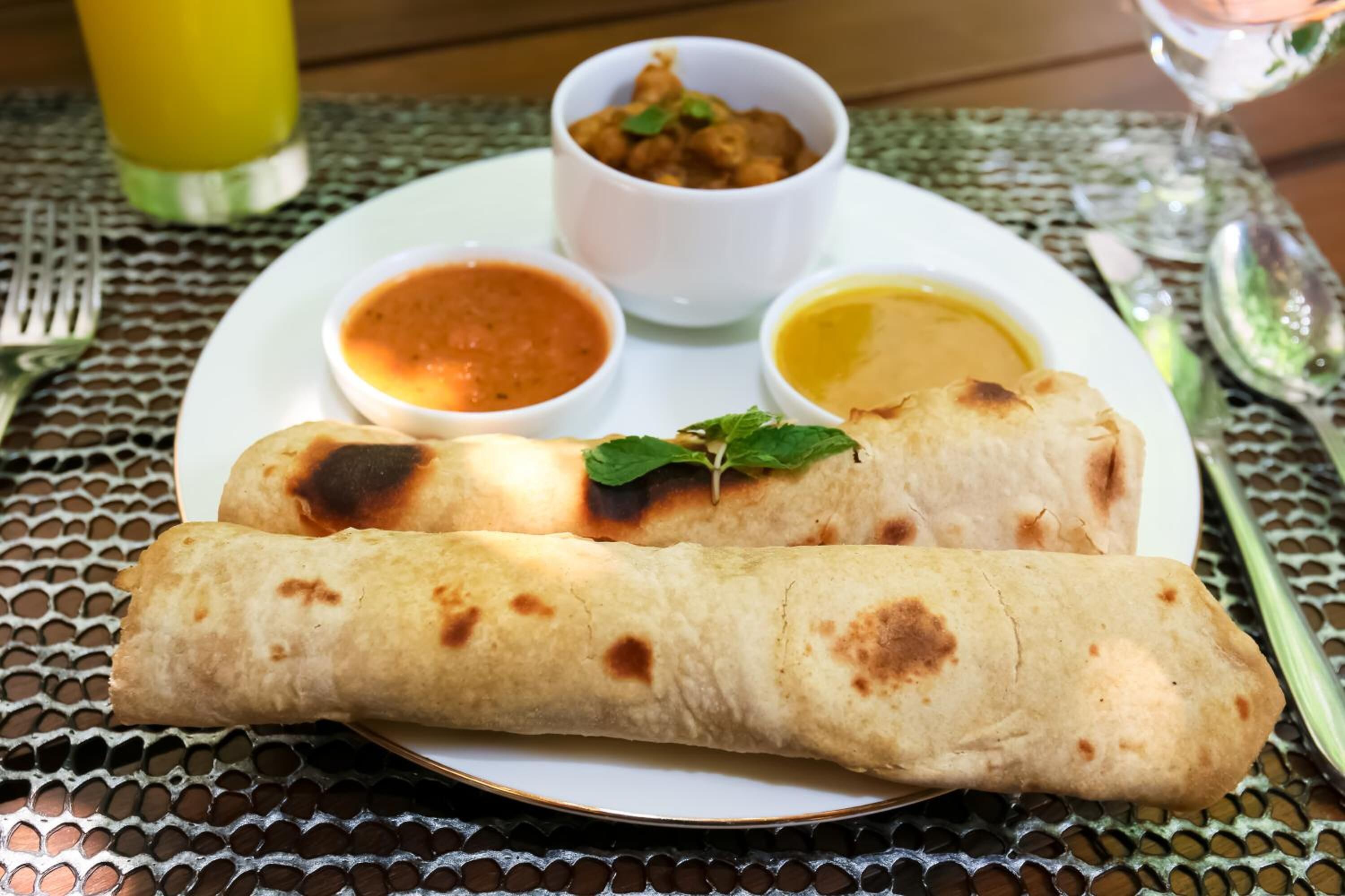 A plate with two rolled flatbreads, two small bowls of sauce, and a small bowl of fried food, all on a table with a woven tablecloth.