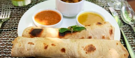 A plate with two rolled flatbreads, two small bowls of sauce, and a small bowl of fried food, all on a table with a woven tablecloth.