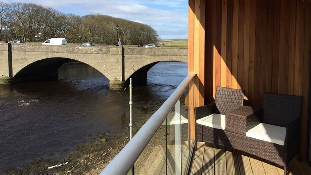 A wooden deck with wicker furniture overlooking a river and a bridge. - Riverside Apartments (Wick)