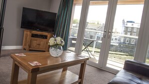 A living room with a wooden table, a flat-screen TV on a wooden stand, a vase with flowers, and a view of buildings through large windows. - Riverside Apartments (Wick)