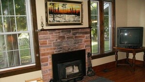 Una sala de estar con chimenea de piedra, un televisor sobre un pedestal de madera y ventanas amplias con persianas.