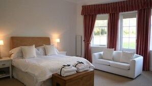Um quarto com cama, mesas de cabeceira, um baú de madeira, um sofá branco e uma janela com cortinas.
