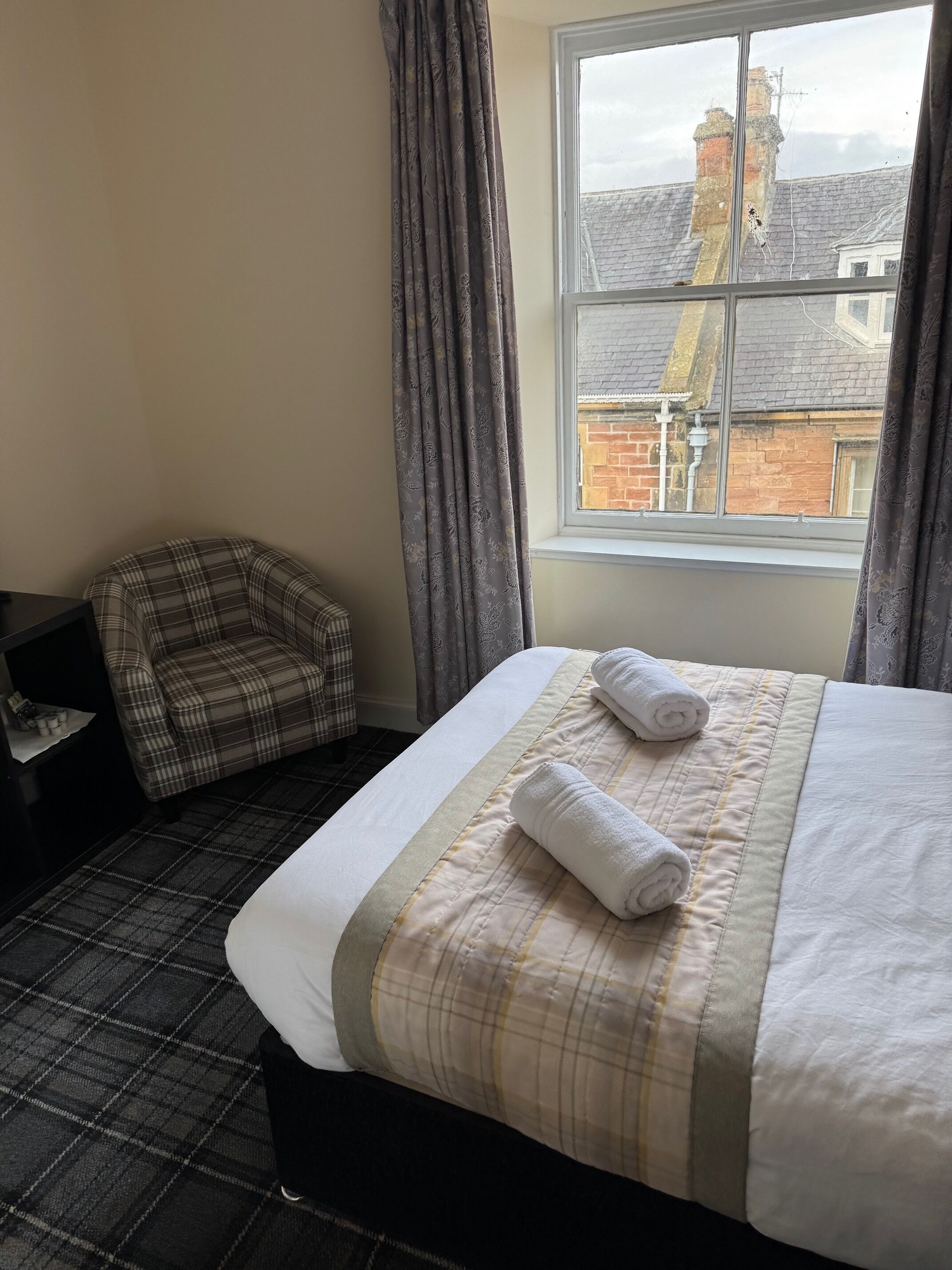 A hotel room with a bed, a plaid armchair, and a window with a view of buildings.