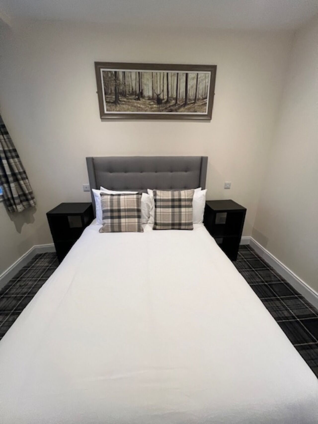 A neatly made bed with a grey headboard, two bedside tables, and a framed picture of a forest on the wall.