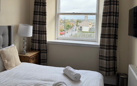 A hotel room with a bed, a nightstand with a lamp, a window with curtains, and a view of the city.