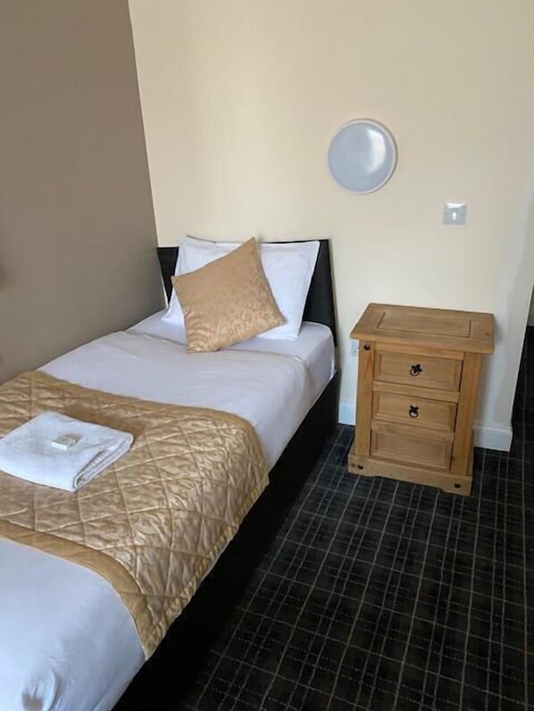 A neatly made bed with a beige quilt and a white pillow, a small wooden nightstand with drawers, and a wall-mounted light fixture.