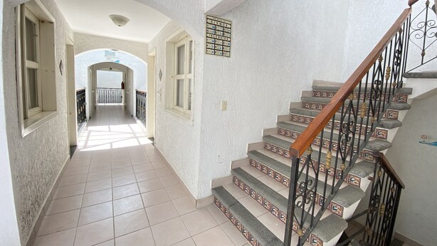 Un pasillo con una escalera que conduce a un piso superior. El pasillo tiene suelo de baldosas y paredes blancas con textura. Hay ventanas a ambos lados del pasillo y un letrero sobre la escalera.