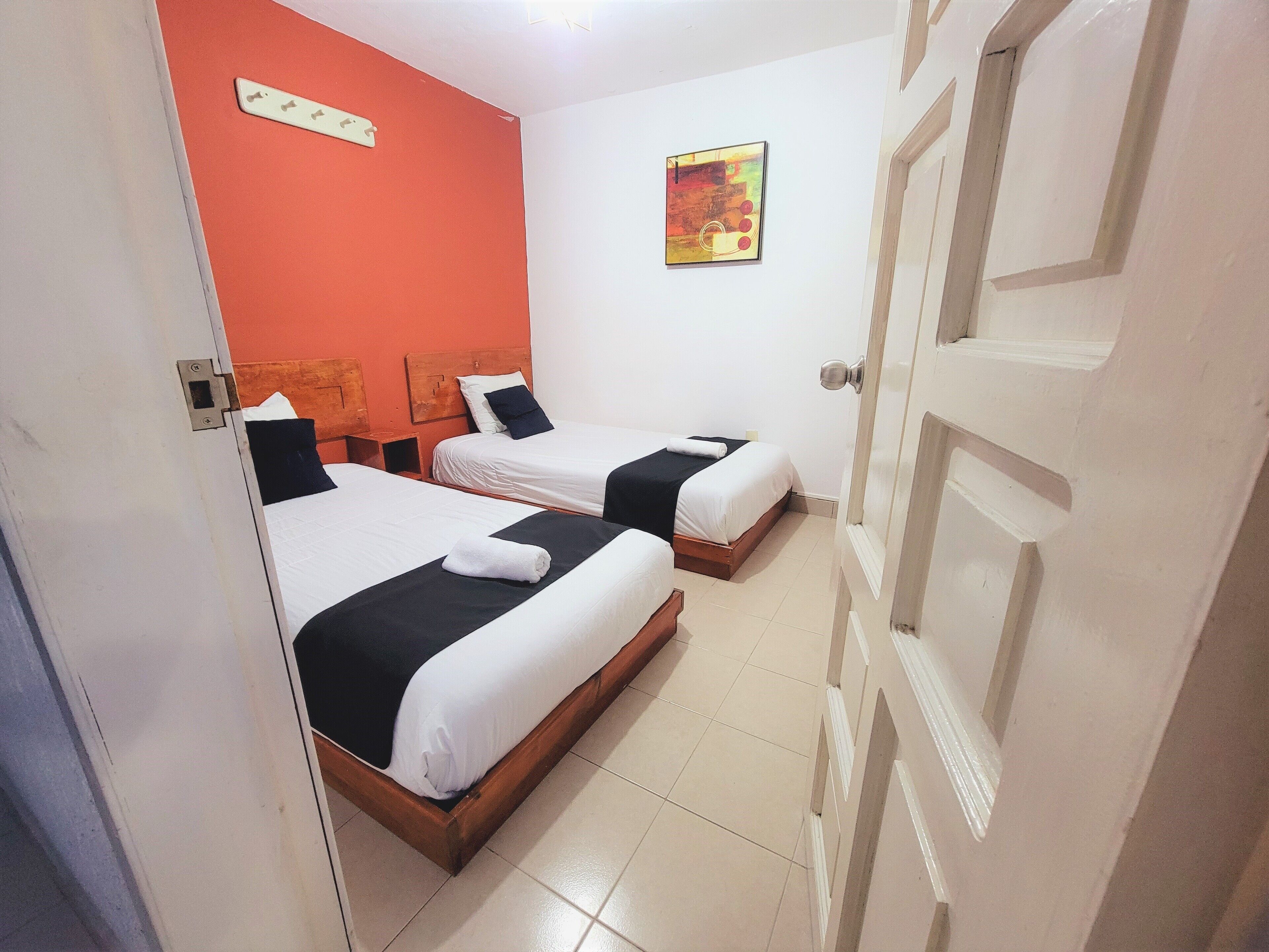 A hotel room with two beds, a red accent wall, a painting, and a white door.