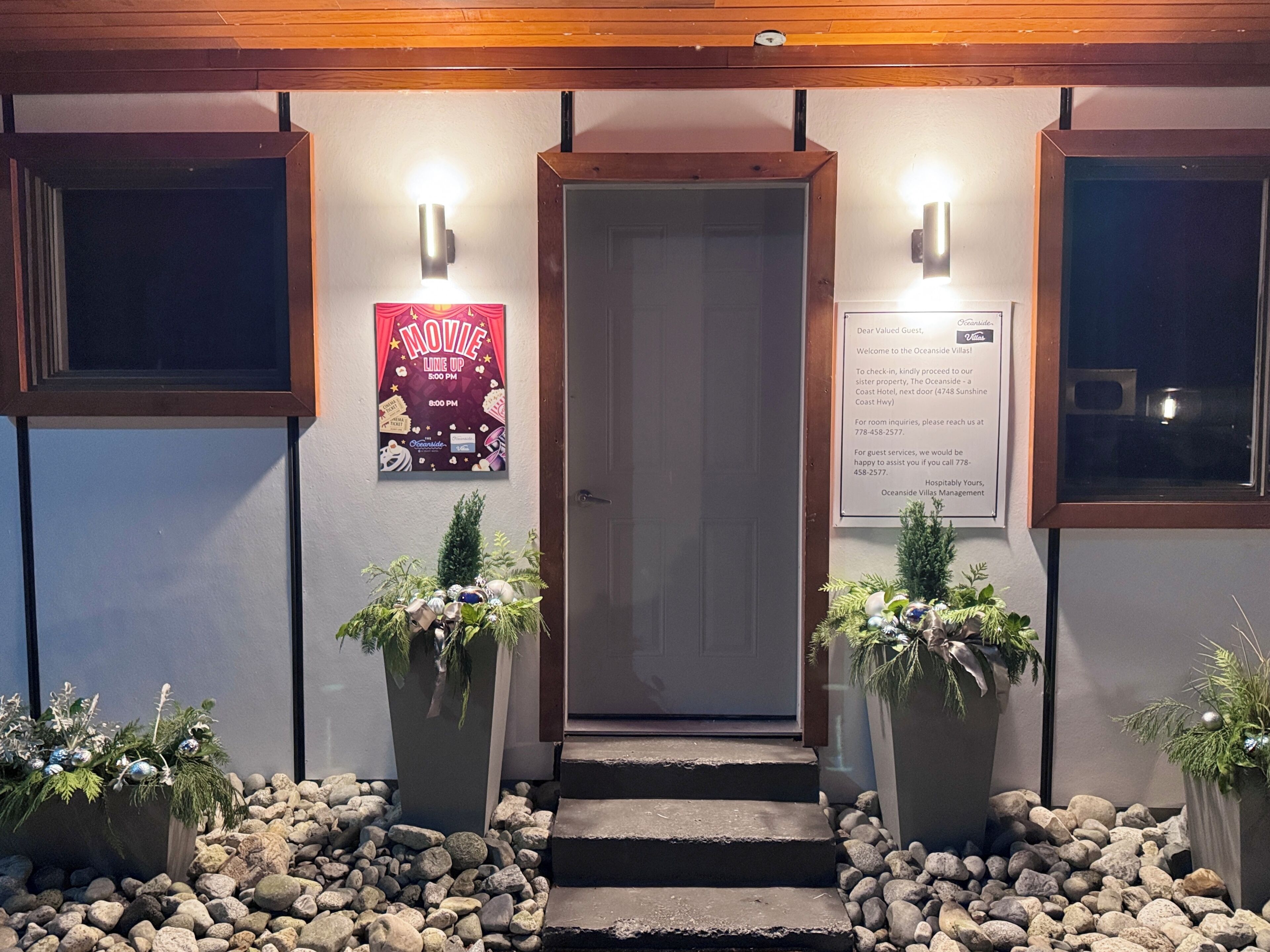 L’entrée d’un hôtel, avec une porte en bois, deux plantes en pot, une affiche et un panneau indicateur.