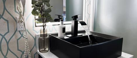A bathroom with a black sink, a hairdryer on the wall, and a plant in a glass vase.