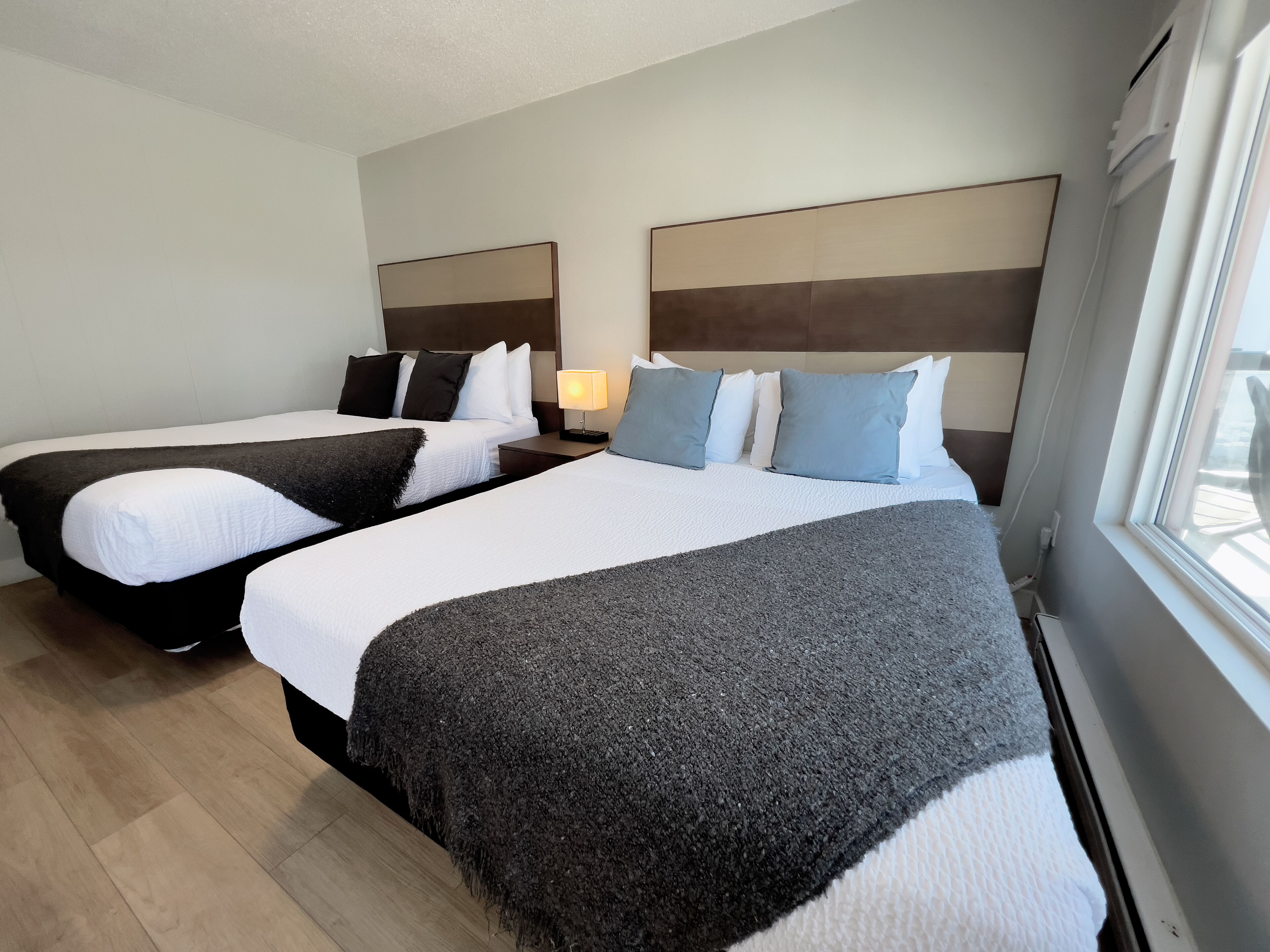 A hotel room with two beds, a nightstand, a lamp, and a window.