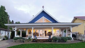 A two-story house with a prominent gable roof, a front porch with a bench, and a large potted plant in the center.