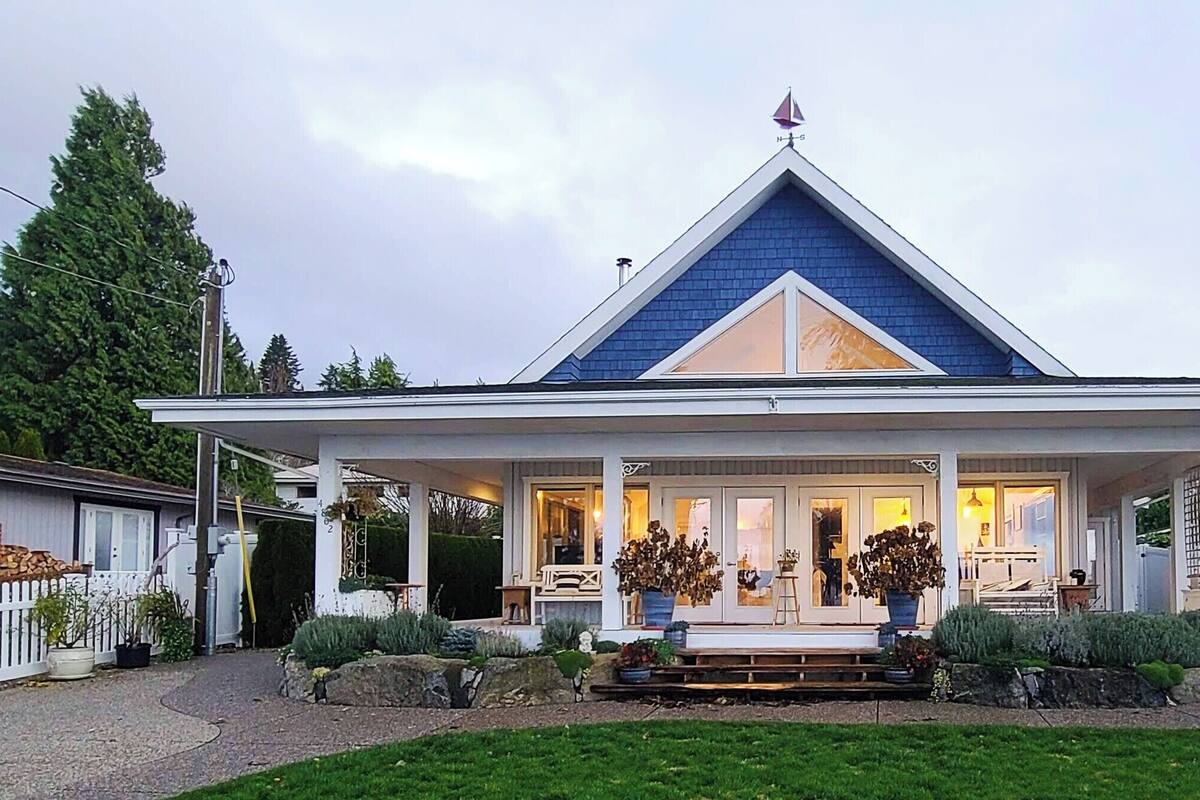 A two-story house with a prominent gable roof, a front porch with a bench, and a large potted plant in the center.