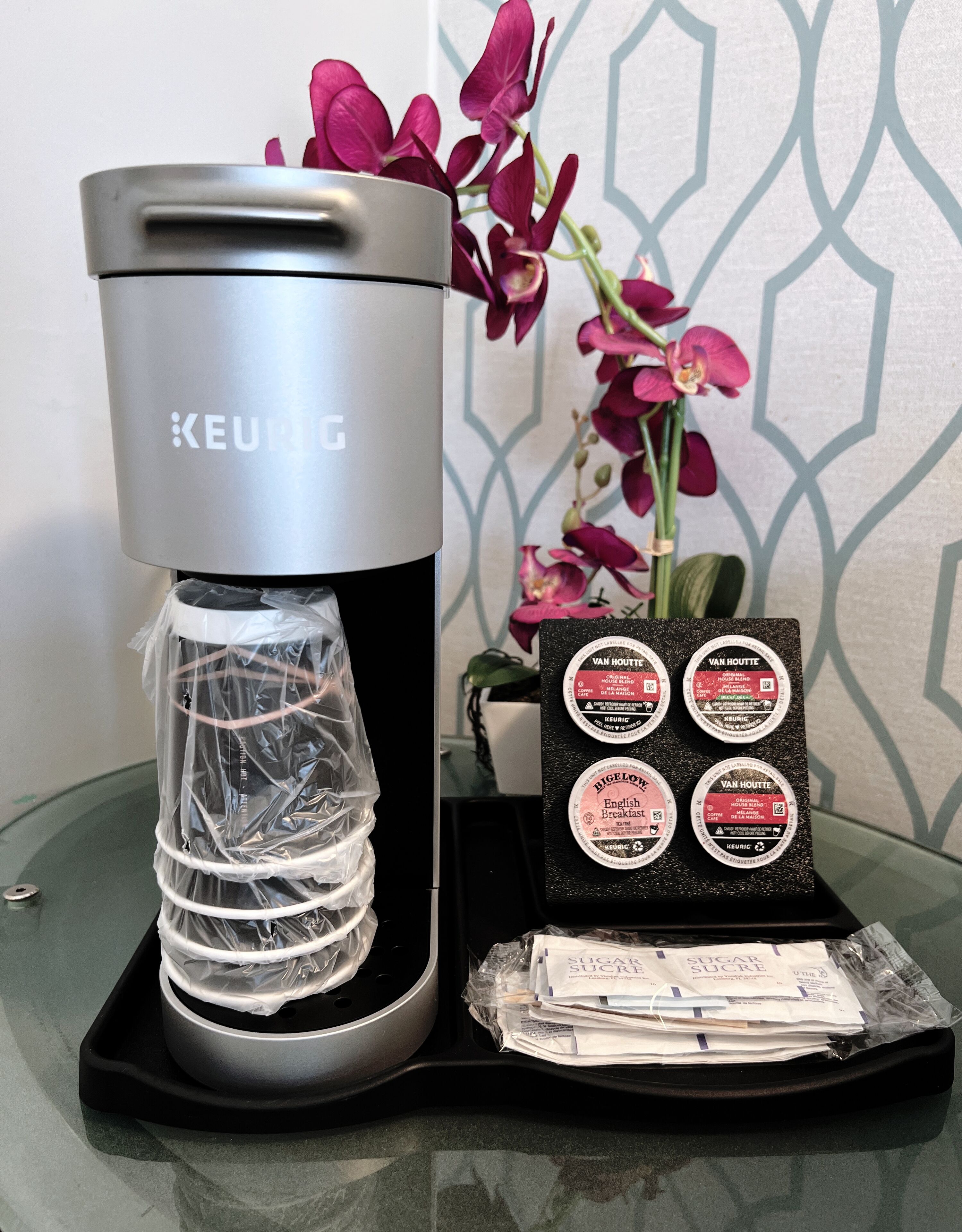 A Keurig coffee maker with a water reservoir, a coffee pod holder, and a stack of sugar packets on a glass table.
