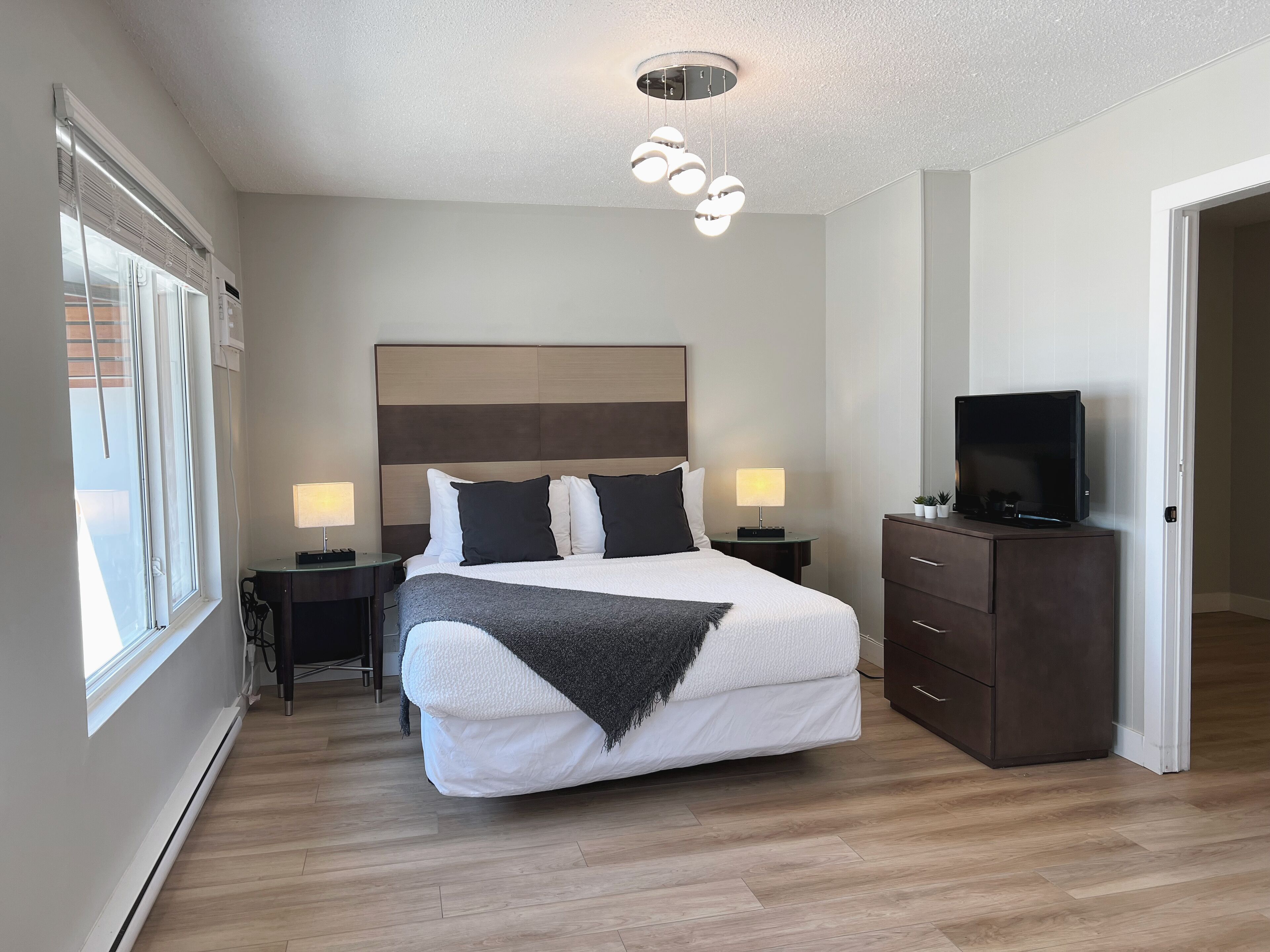 A modern hotel room with a large bed, bedside lamps, a flat-screen TV, and a wooden floor.