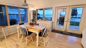 A dining area with a wooden table and four chairs, large windows with a view of the sea, a white cabinet, and a small shelf with decorative items.
