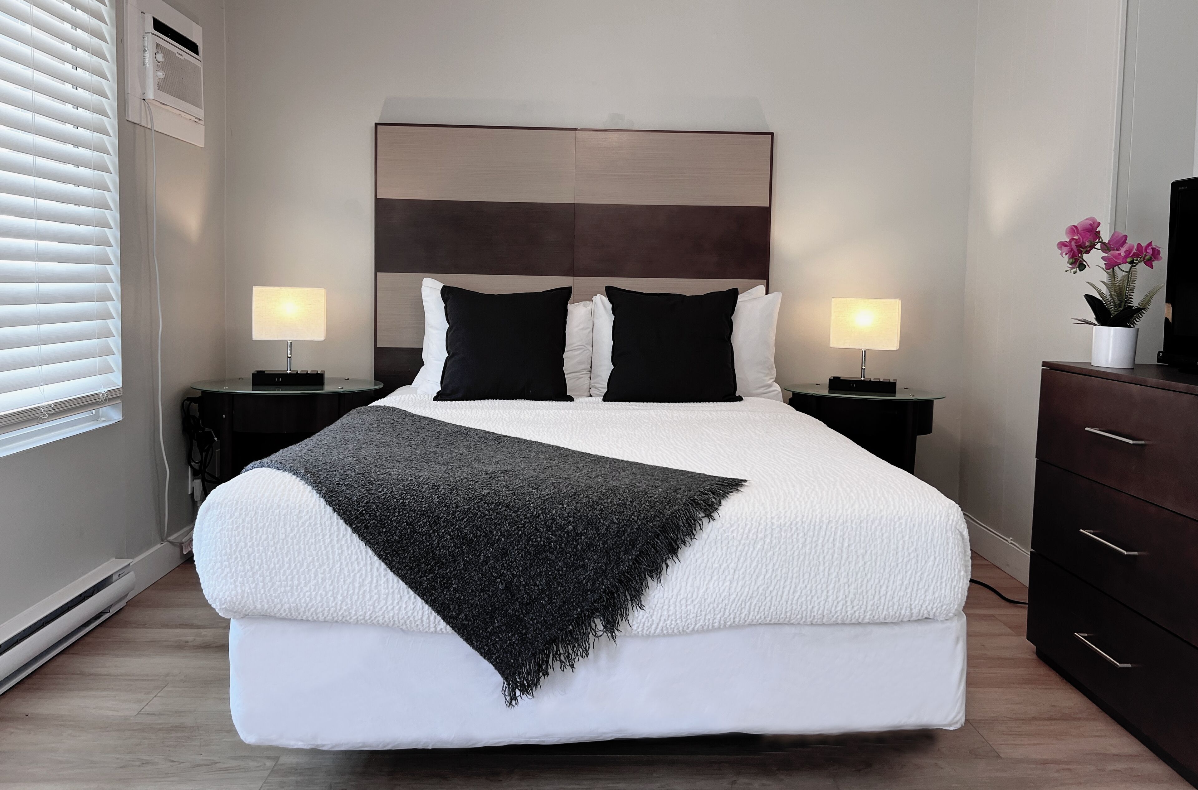 Habitación de hotel con una cama grande, dos mesitas de noche con lámparas, un cabecero de madera oscura, una manta negra y una planta en maceta sobre un aparador.
