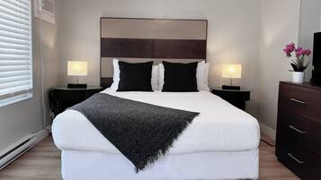Habitación de hotel con una cama grande, dos mesitas de noche con lámparas, un cabecero de madera oscura, una manta negra y una planta en maceta sobre un aparador.