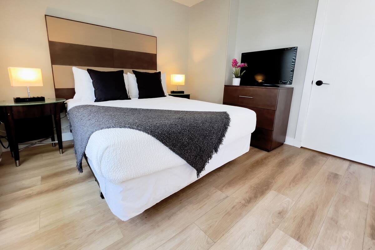 A neatly arranged hotel room with a large bed, bedside tables with lamps, a dresser, a television, and a door.
