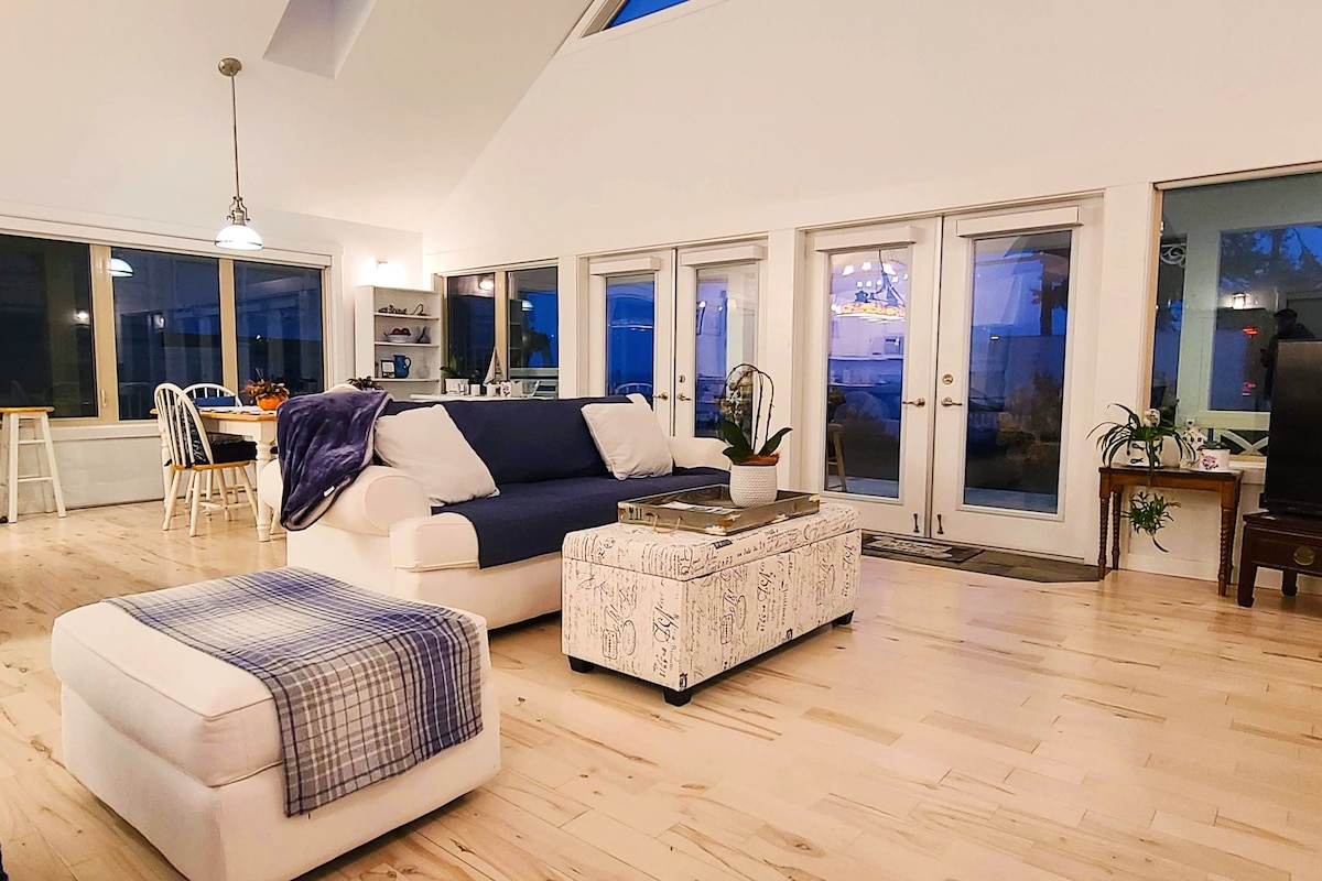 A spacious living room with a large sofa, a coffee table, and a TV. There is a dining area with a table and chairs, and a kitchen in the background.