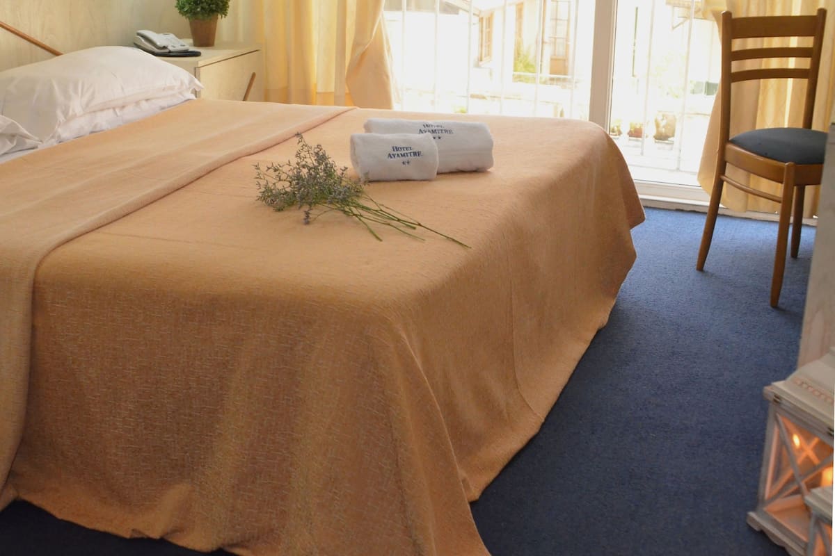 A neatly made bed with a beige bedspread, two rolled towels with text, a small plant, and a chair with a cushion.