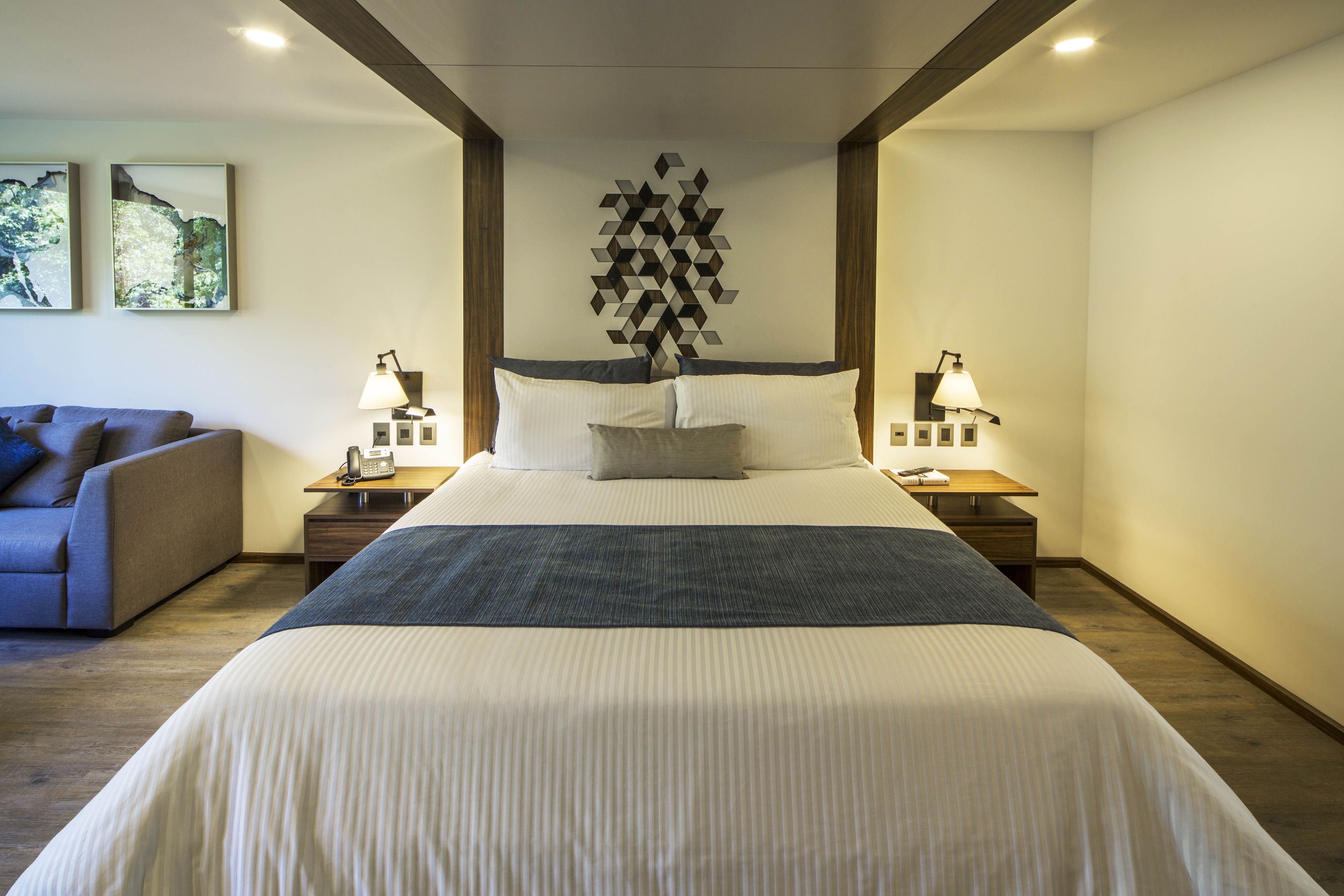 Un cuarto de hotel ordenado con una cama grande, dos mesitas de noche con lámparas, un sofá y arte abstracto en la pared.
