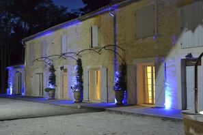A building with multiple doors and windows, some with shutters, and a paved entrance area. - Le 7 Mas Provençal (Saint-Andiol)