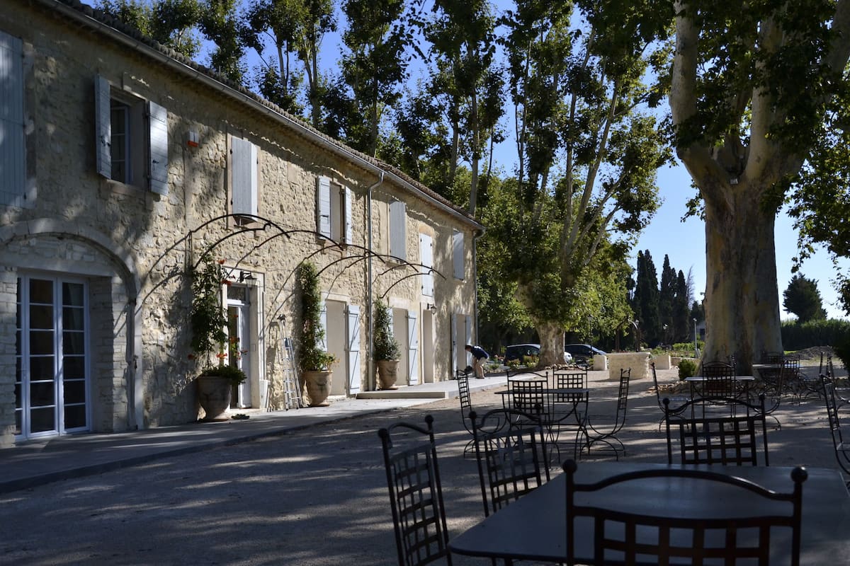 A stone building with a row of windows and shutters, outdoor seating with metal chairs and tables, and a large tree in the background.