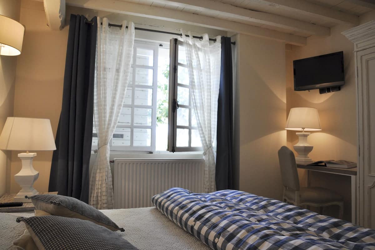 A bedroom with a bed, two bedside lamps, a chair, a desk with a lamp, a television on the wall, and a window with curtains.