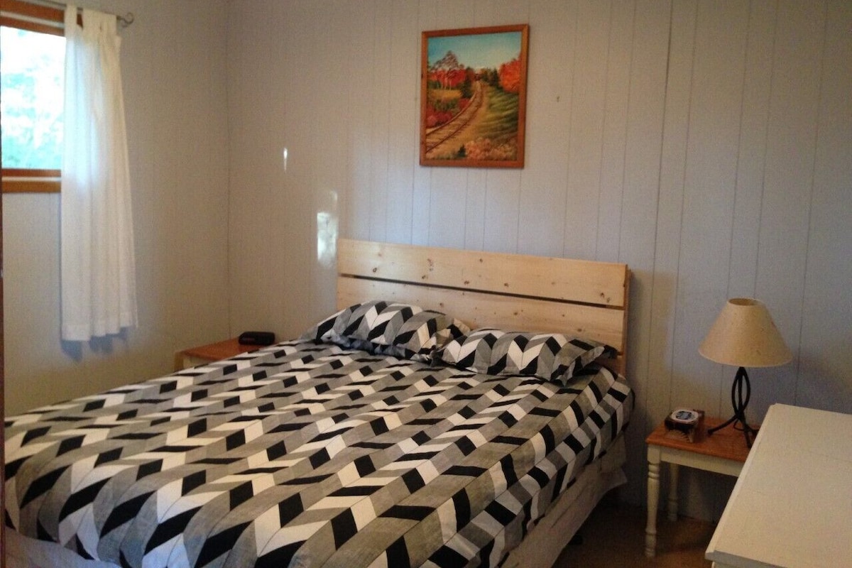 Un dormitorio con cabecera de madera, cama con colcha estampada, mesita de noche con lámpara y un cuadro en la pared.