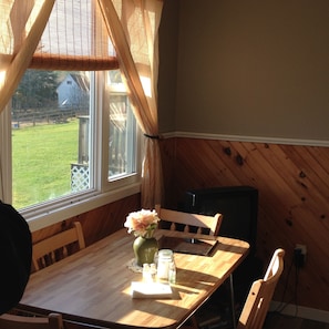 Una mesa de comedor de madera con un florero, saleros y pimenteros, y una servilleta. Se ve una ventana con cortinas que ofrece vistas a un patio cubierto de césped y un edificio.