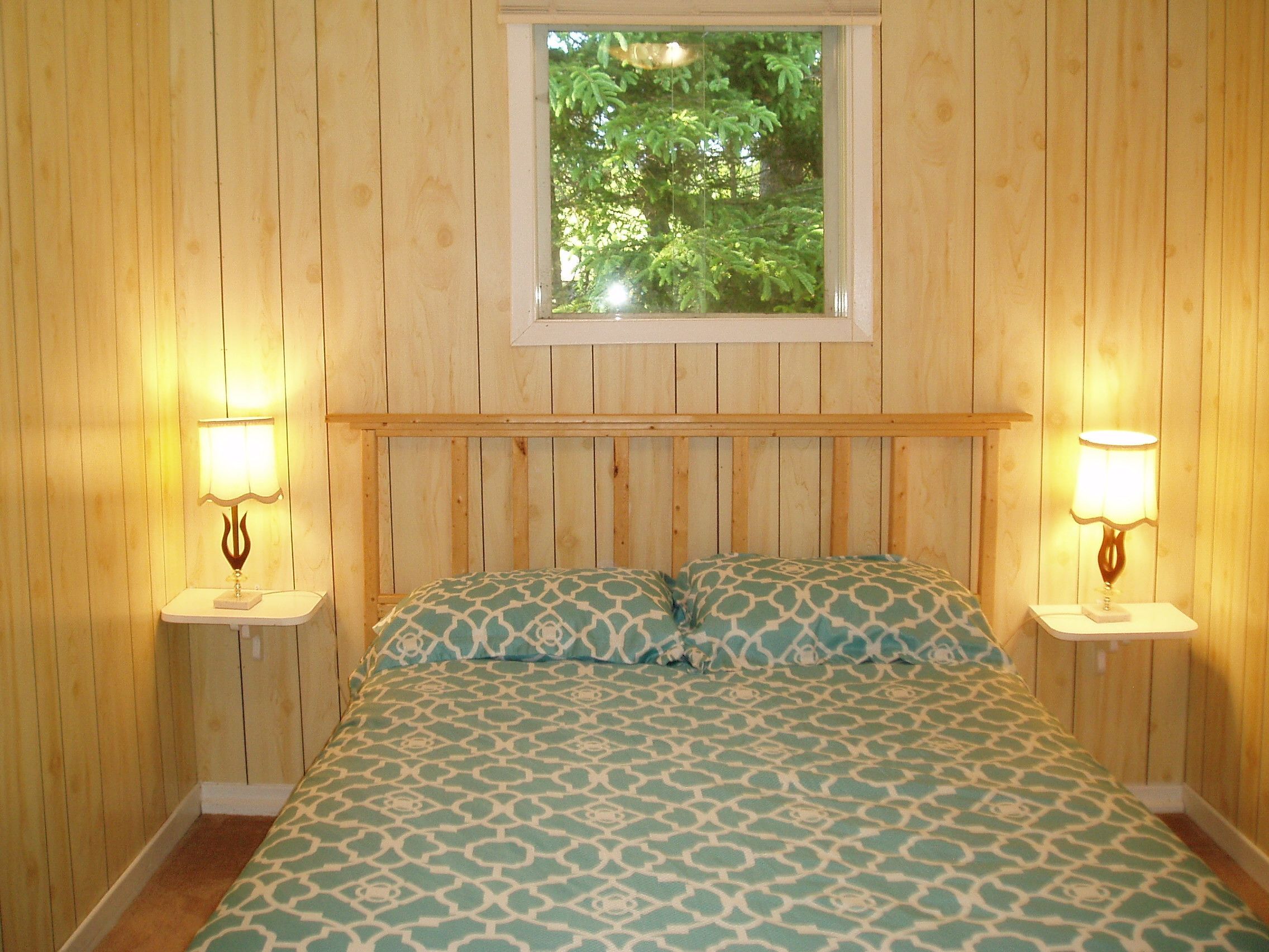 Um quarto com painéis de madeira, uma cama, duas mesas de cabeceira com abajures e uma janela com vista para árvores.