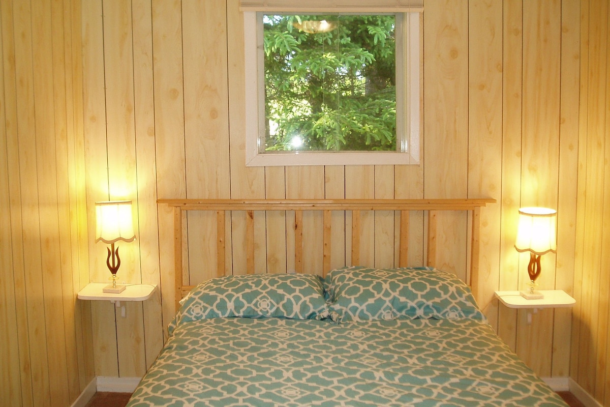 Una habitación con paneles de madera, una cama, dos mesitas de noche con lámparas y un ventanal con vistas a árboles.