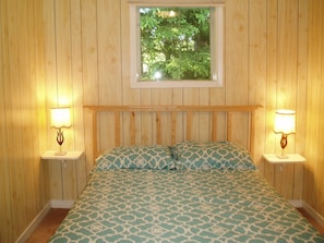 Una habitación con paneles de madera, una cama, dos mesitas de noche con lámparas y un ventanal con vistas a árboles.
