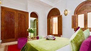 A room with a bed, a wooden wardrobe, a small table with a vase of flowers, and a purple ottoman. - Dar Pamella (Marrakech)