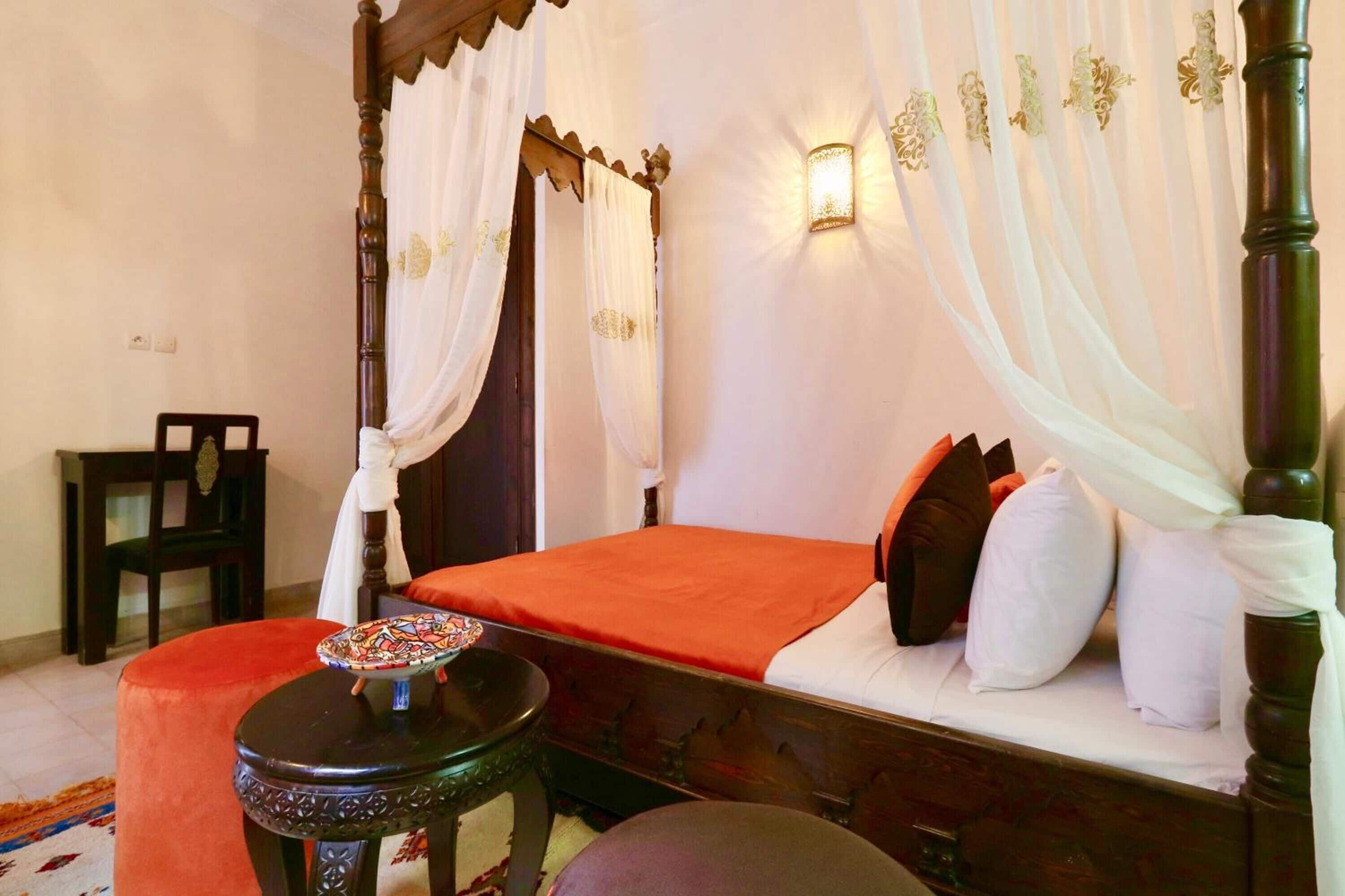 A four-poster bed with white drapes, a red bedspread, and white pillows. There is a small round table with a colorful object on it.