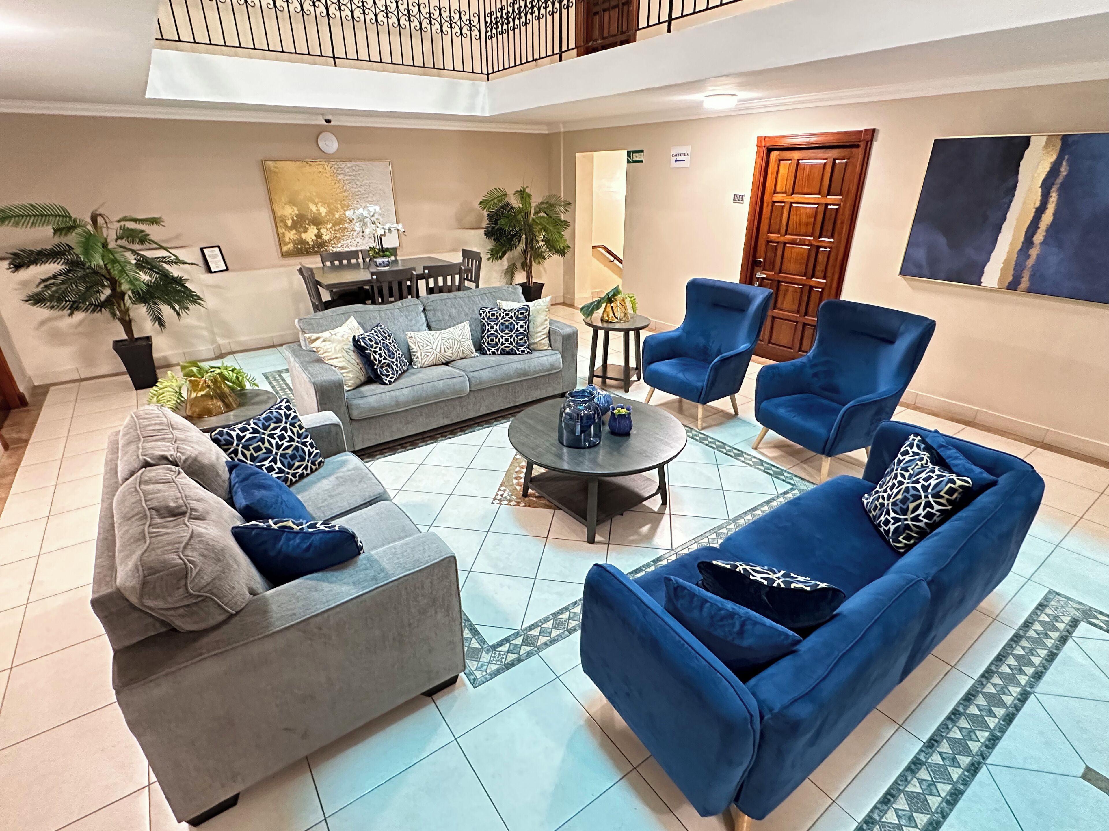 Un vaste espace de vie comprenant un canapé gris, des fauteuils bleus et une table basse. Au fond, on aperçoit une table à manger avec des chaises, et un escalier menant à l’étage supérieur.