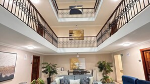 A spacious living area with a central coffee table, two sofas, and a blue armchair. A dining table is set with chairs. The room features a high ceiling with a skylight and a balcony with wrought iron railings. - Hotel Andros (Colon)
