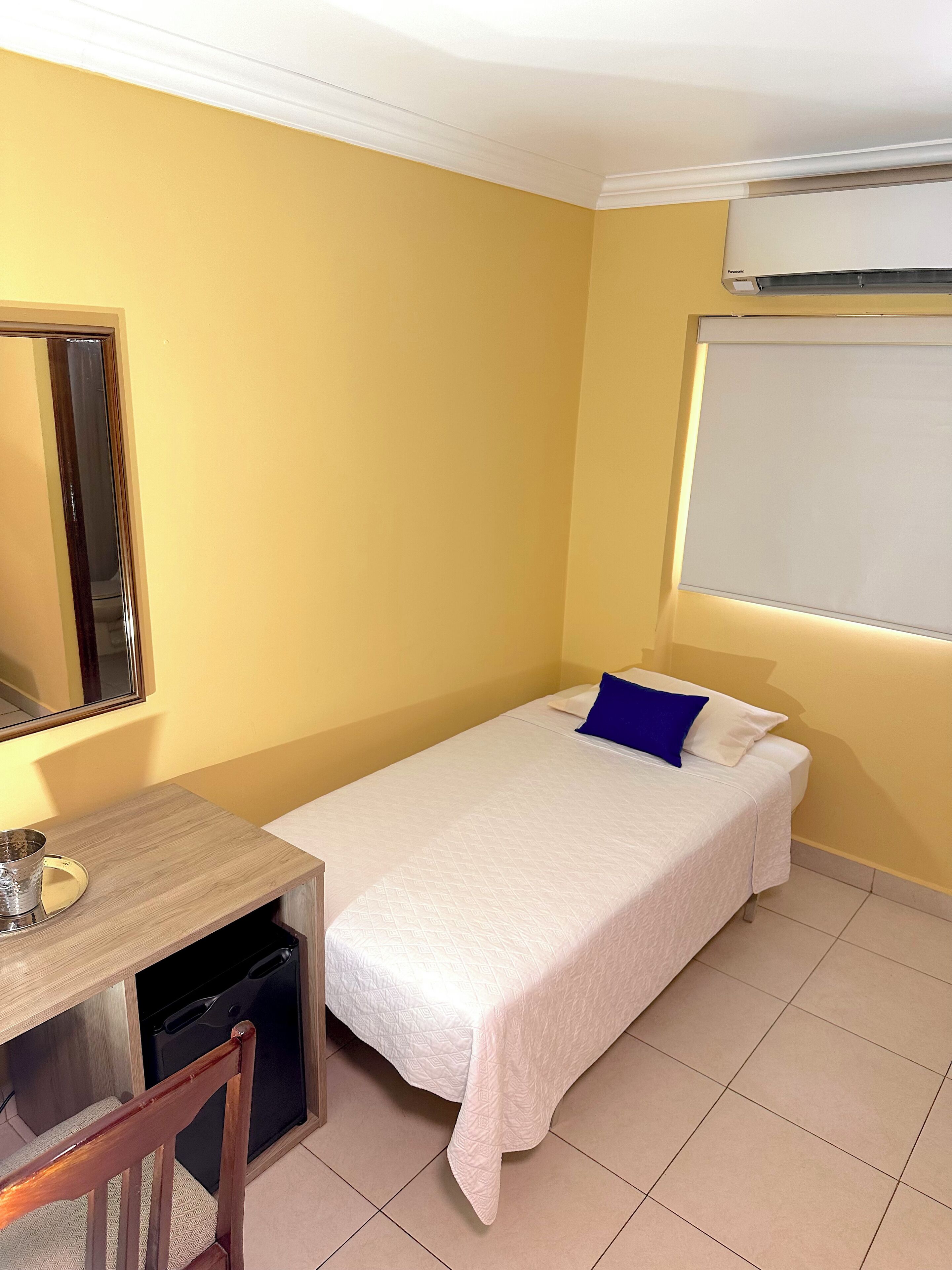 Una habitación pequeña con cama individual, escritorio, silla, espejo y ventana con unidad de aire acondicionado.