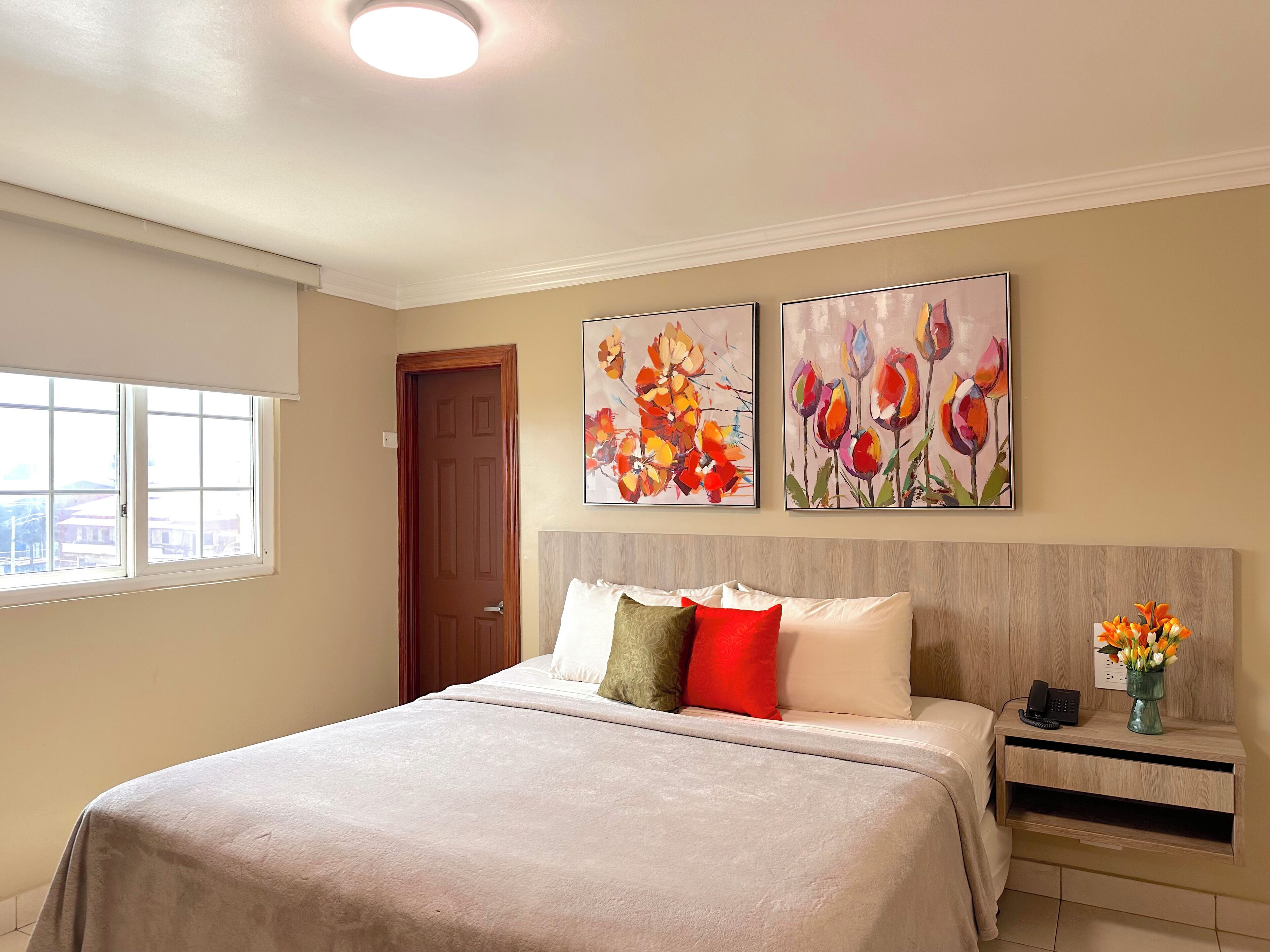 A bedroom with a bed, a nightstand, a window, and two floral paintings on the wall.