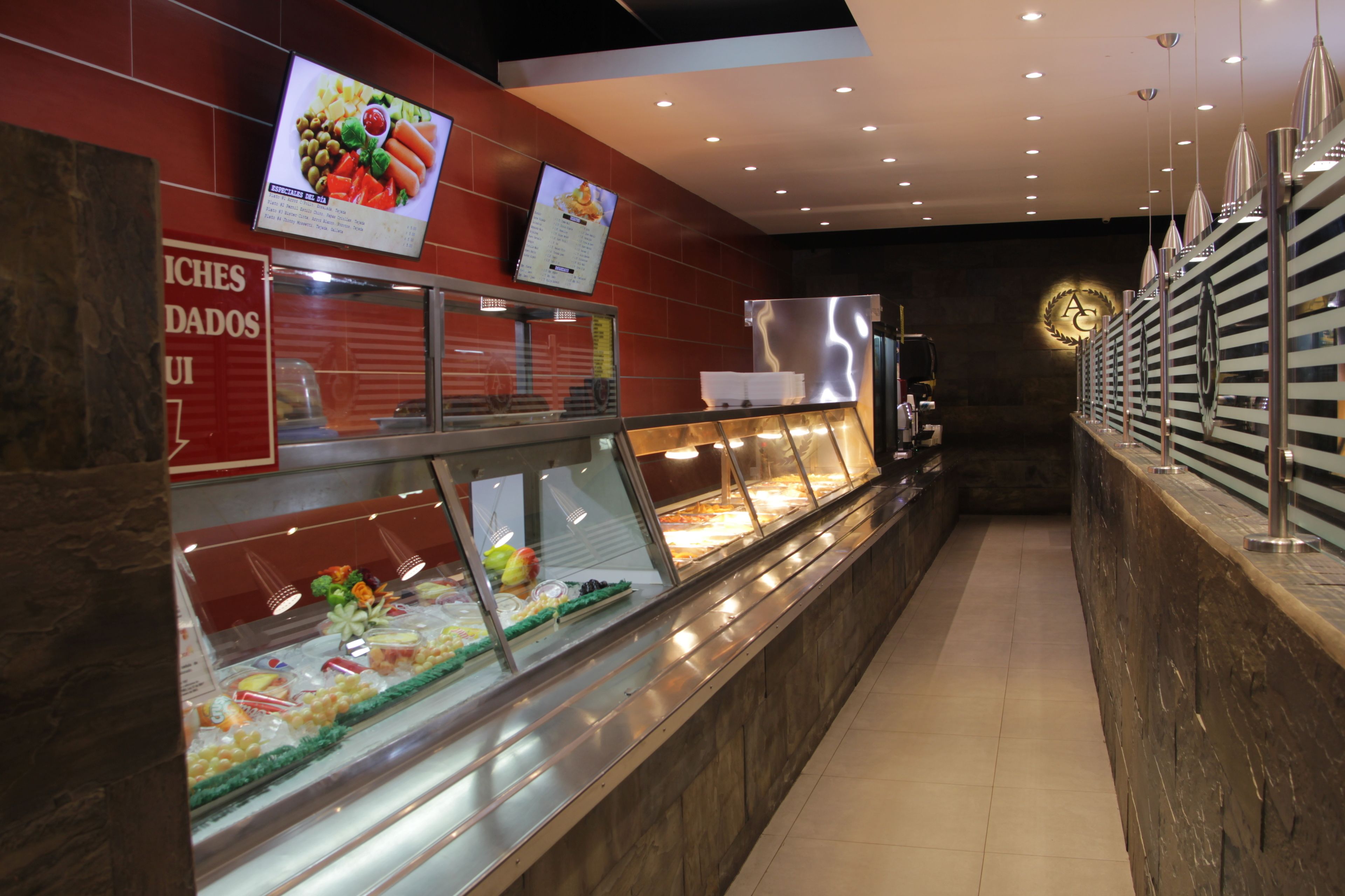 Zona de bufé con un expositor de vidrio lleno de diferentes alimentos, un menú digital arriba y un refrigerador de acero inoxidable.