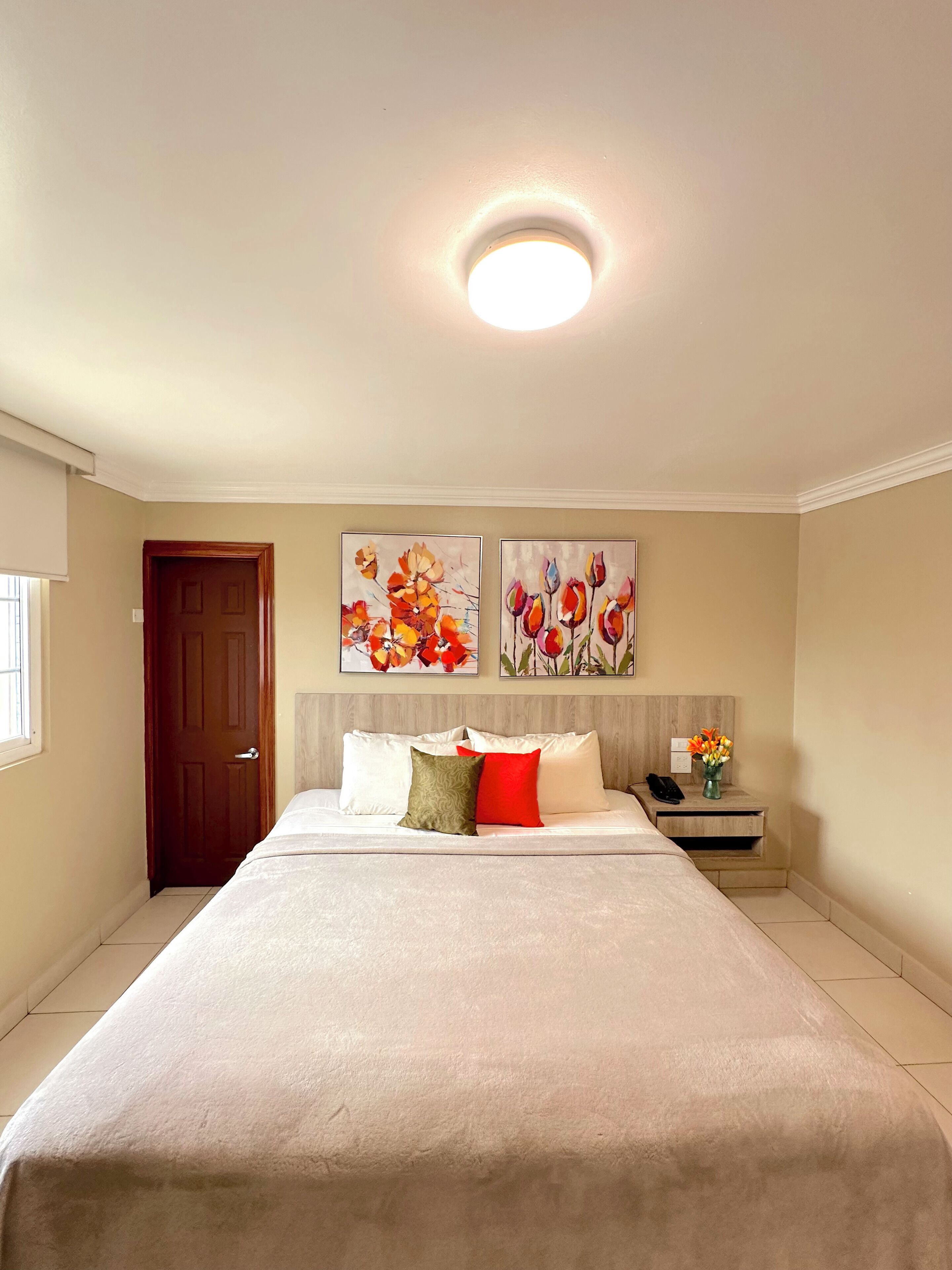 A hotel room with a large bed, a wooden headboard, two wall-mounted paintings, a bedside table with a phone and a vase of flowers, and a door with a window.