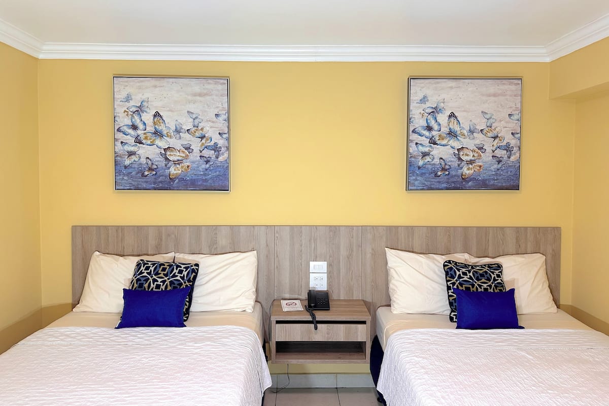 A hotel room with two beds, each with white bedding and patterned pillows. There are two paintings of butterflies on the yellow walls.