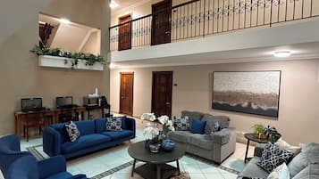 Amplia sala de estar con dos sofás, una mesa de centro y un televisor. Hay un balcón con barandilla y puerta.