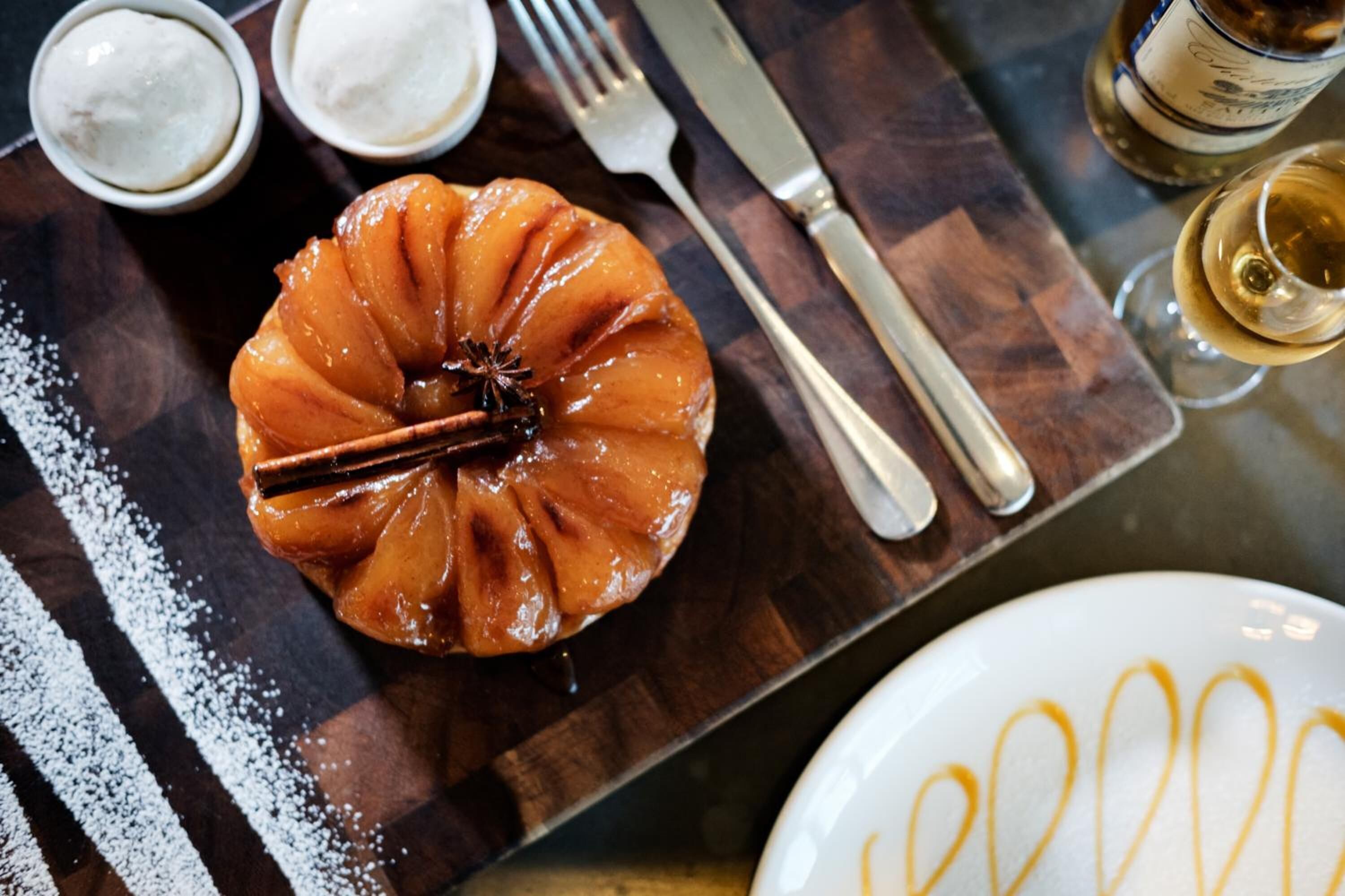 A caramel-glazed apple dessert on a wooden board, accompanied by a fork, knife, and a glass of white wine.