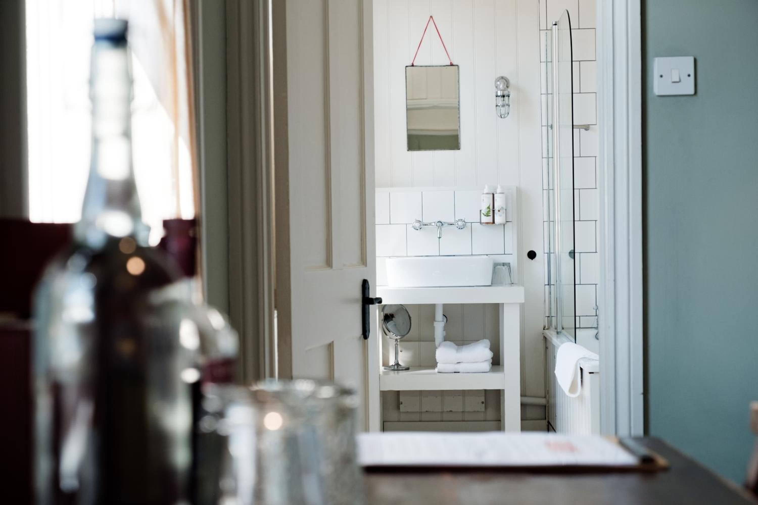 Un baño con lavamanos blanco, espejo y toallero. Hay una botella y un vaso sobre el mostrador.